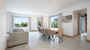 Ein geräumiger Wohnbereich mit Esstisch und Stühlen, große Fenster mit Blick auf Grünflächen und ein modernes abstraktes Gemälde an der Wand.