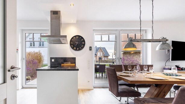 A modern kitchen with a central island, a dining area with a wooden table and chairs, and a large window offering a view of a house. - Holiday Home 'Treibholz' with Private Terrace, Private Garden and Wi-Fi (Dassow)
