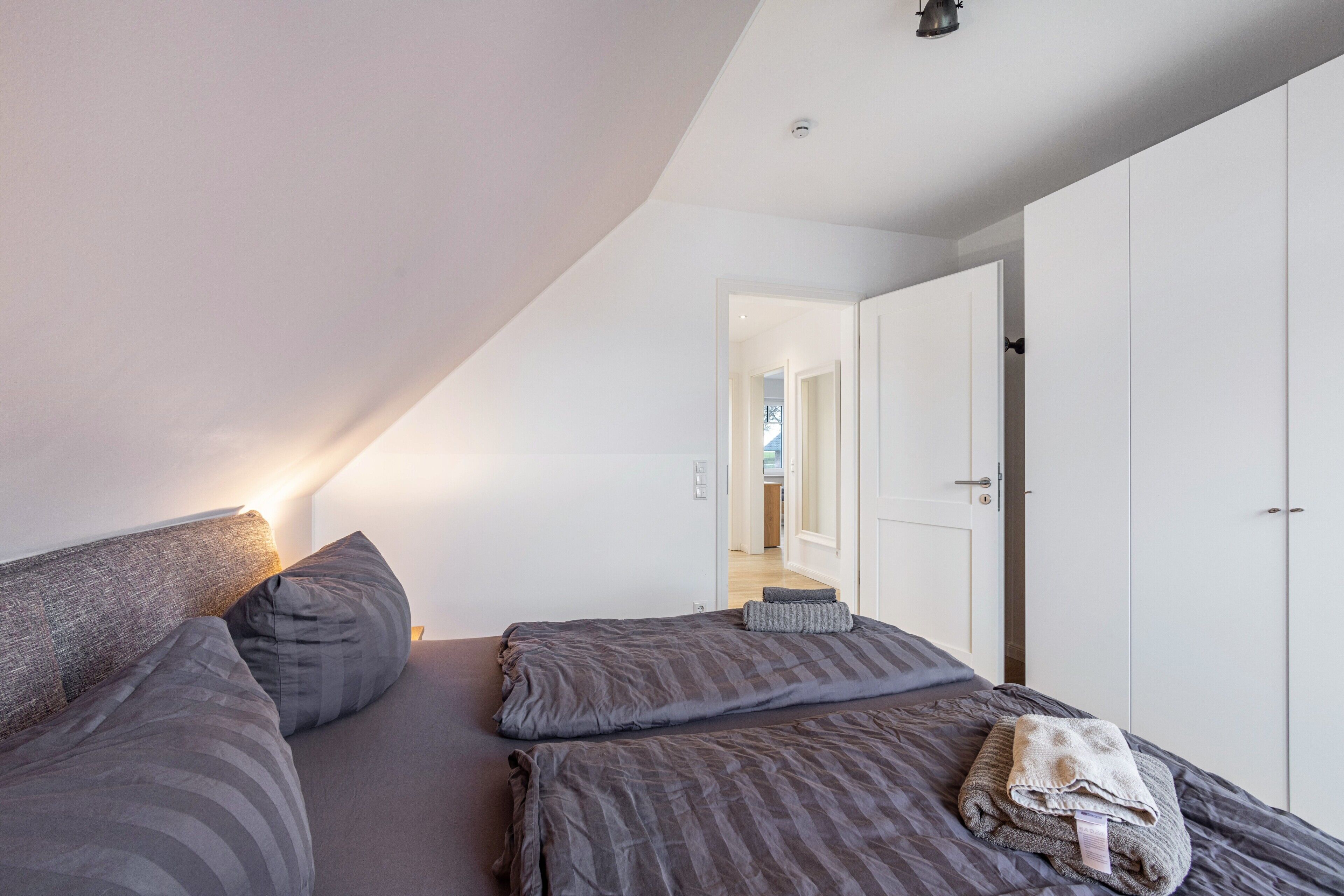 Un dormitorio con techo inclinado, una cama con ropa de cama gris, una lámpara de noche y un armario.