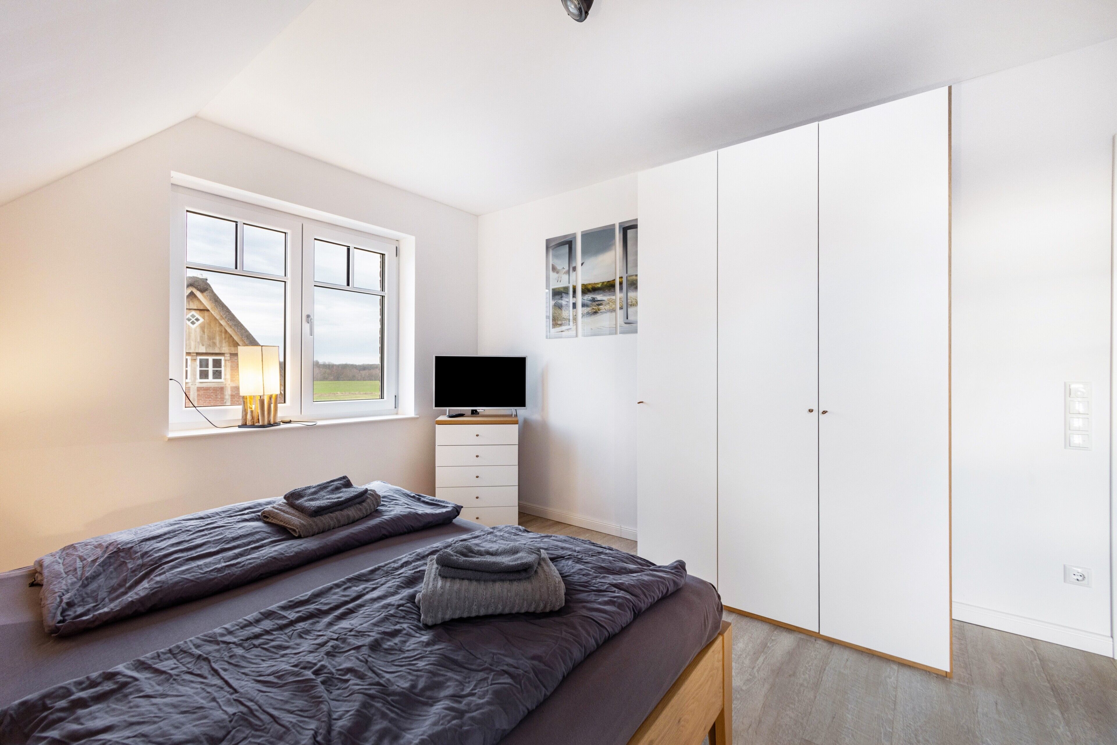 A bedroom with a bed, a TV, a wardrobe, and a window with a view of a house.