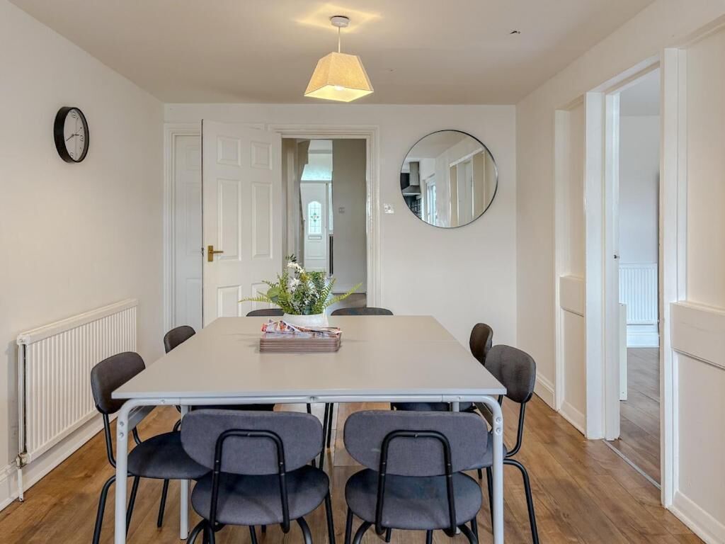 Een eethoek met een witte tafel, zes stoelen, een hanglamp, een ronde spiegel en een klok aan de muur.