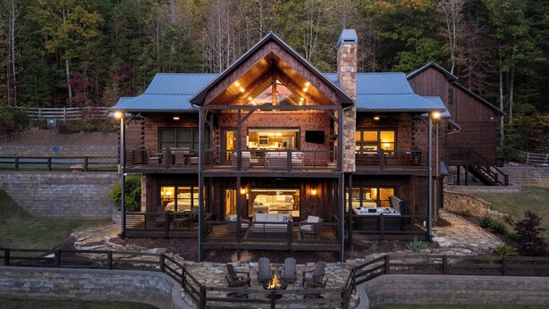 A large, multi-story log cabin with a prominent chimney, a balcony with outdoor seating, and a stone fire pit in the foreground. - Great Trails Lodge - Blue Ridge (Epworth)