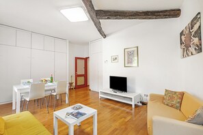 En moderne stue med spiseplads, trægulv, et fjernsyn på et hvidt skab og en gul sofa.