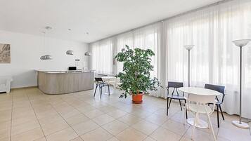Ein modernes Hotelzimmer mit Küchenzeile, einem Esstisch, Stühlen, einer Kübelpflanze und großen Fenstern mit durchsichtigen Vorhängen.