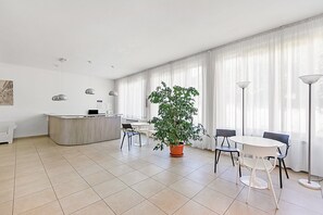 A modern hotel room with a kitchen area, a dining table, chairs, a potted plant, and large windows with sheer curtains. - Bogliaco Lake Front (Gargnano)