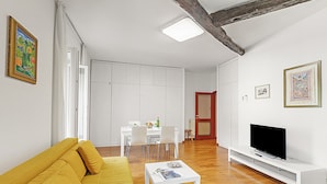 Un salon comprenant un canapé, un téléviseur posé sur un meuble, un coin repas avec des chaises et un buffet blanc.