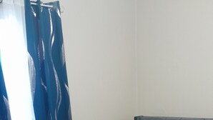 Ein ordentlich bezogenes Bett mit grauer Bettwäsche und einem gefalteten weißen Handtuch, ein flauschiger beiger Teppich und ein Fenster mit blauen Vorhängen.