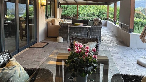 A covered patio with outdoor seating, a glass table, and a view of greenery.