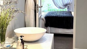 A bathroom with a large mirror, a white sink, and a vase of flowers. A robe hangs on a hook, and a bed is visible through an open door.