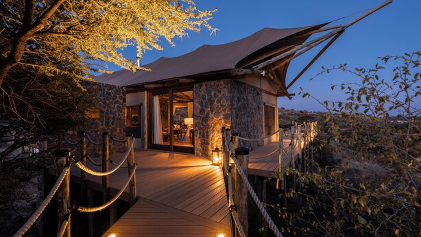 Eine Holzterrasse führt zu einem Steinhaus mit überdachter Eingang, umgeben von Bäumen und blauem Himmel.