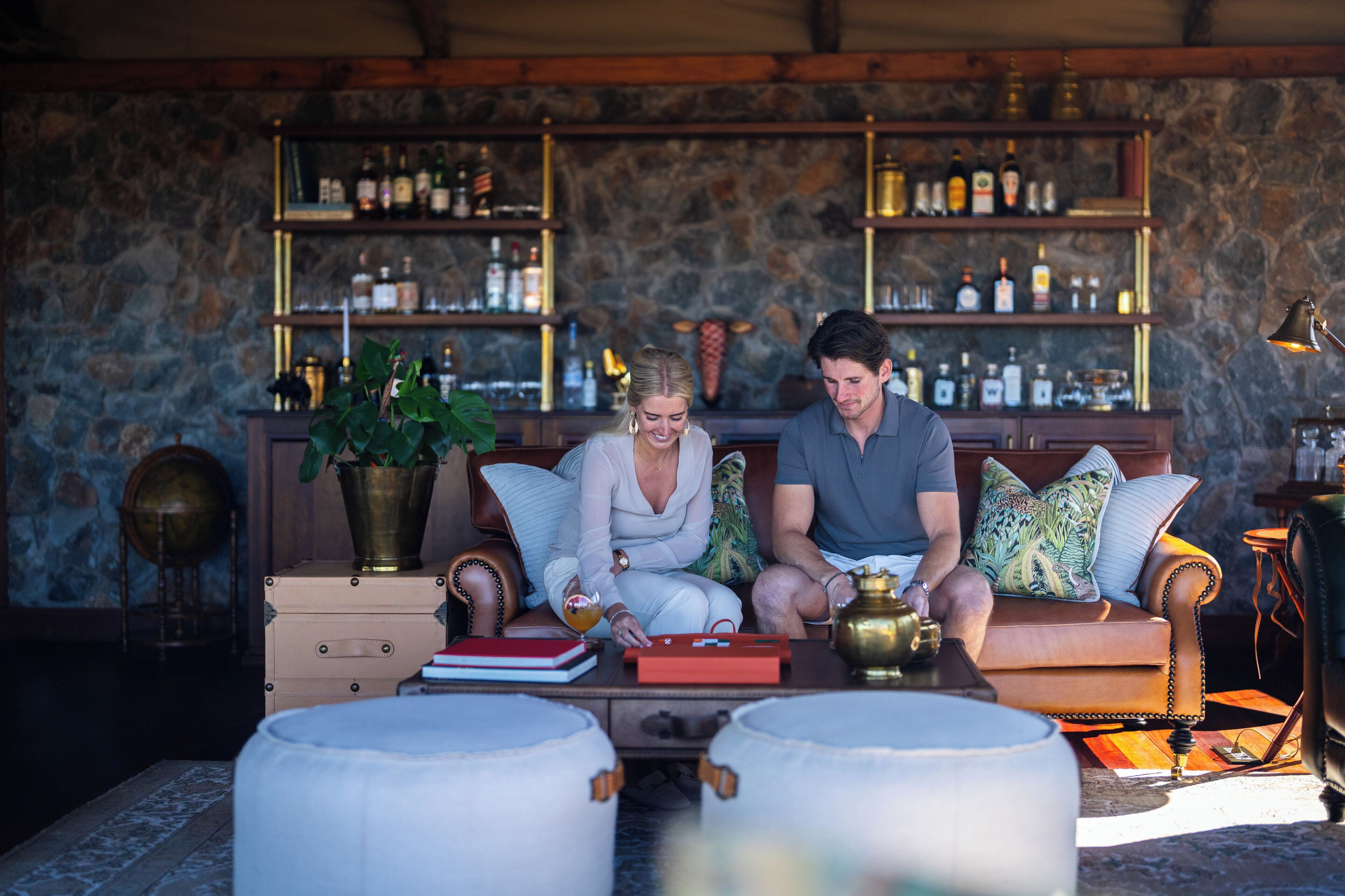 A couple sitting on a sofa in a room with a stone wall and a bar area with shelves of bottles.