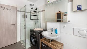 A modern bathroom with a glass shower enclosure, a built-in washing machine, a sink, and a wooden cabinet.
