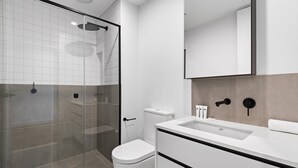 Une salle de bain moderne avec une douche à l’italienne en verre, des toilettes blanches et un grand miroir au-dessus d’un lavabo blanc doté de poignées noires.