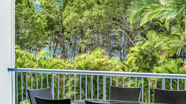 Un balcon agrémenté de fauteuils et d’une table en osier, donnant sur un jardin luxuriant où poussent différentes espèces d’arbres et de plantes.