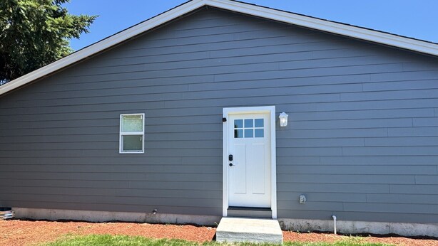 A single-story building with a dark gray exterior, a white front door, and a small window above the door. - Private room and bathroom in safe Vancouver neighborhood (Vancouver)