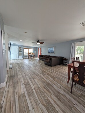 A spacious living area with a large sofa, dining table, and chairs, a ceiling fan, and a view of the outdoors.