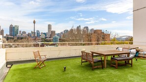A rooftop terrace with a city skyline view, a wooden table and chairs, and a small patch of artificial grass. - Eclectic Art Deco Apartment in Great Location (Sydney)