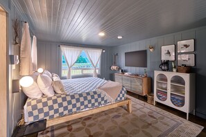 A bedroom with a checkered bedspread, a wooden bench, a dresser with glass doors, and a TV on the wall. - Remodeled | Downtown Long Beach (The Nest) (Long Beach)