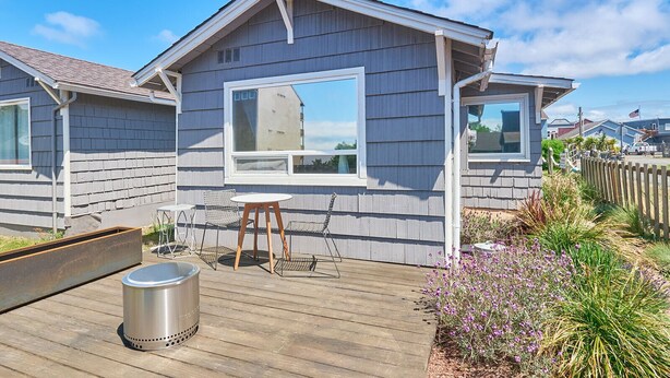 Une maison en bois d’un seul étage, dotée d’une grande fenêtre, d’une petite terrasse et d’une poubelle en métal.
