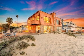 Une propriété située en bord de mer, avec un bâtiment moderne à deux étages, une plage de sable en premier plan et un ciel dégagé.