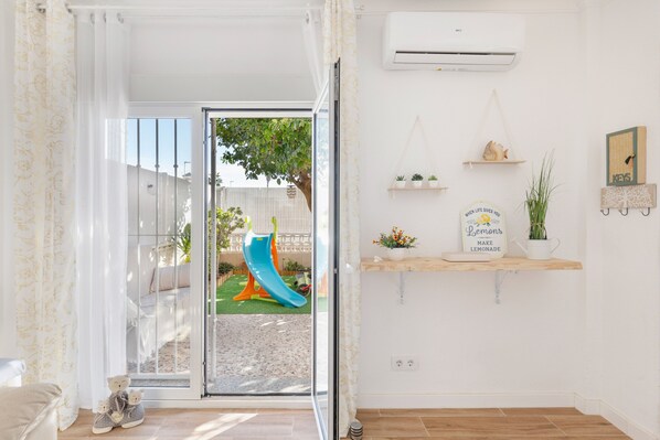 A room with a sliding glass door leading to a backyard with a slide, a white air conditioner on the wall, and a shelf with decorative items.