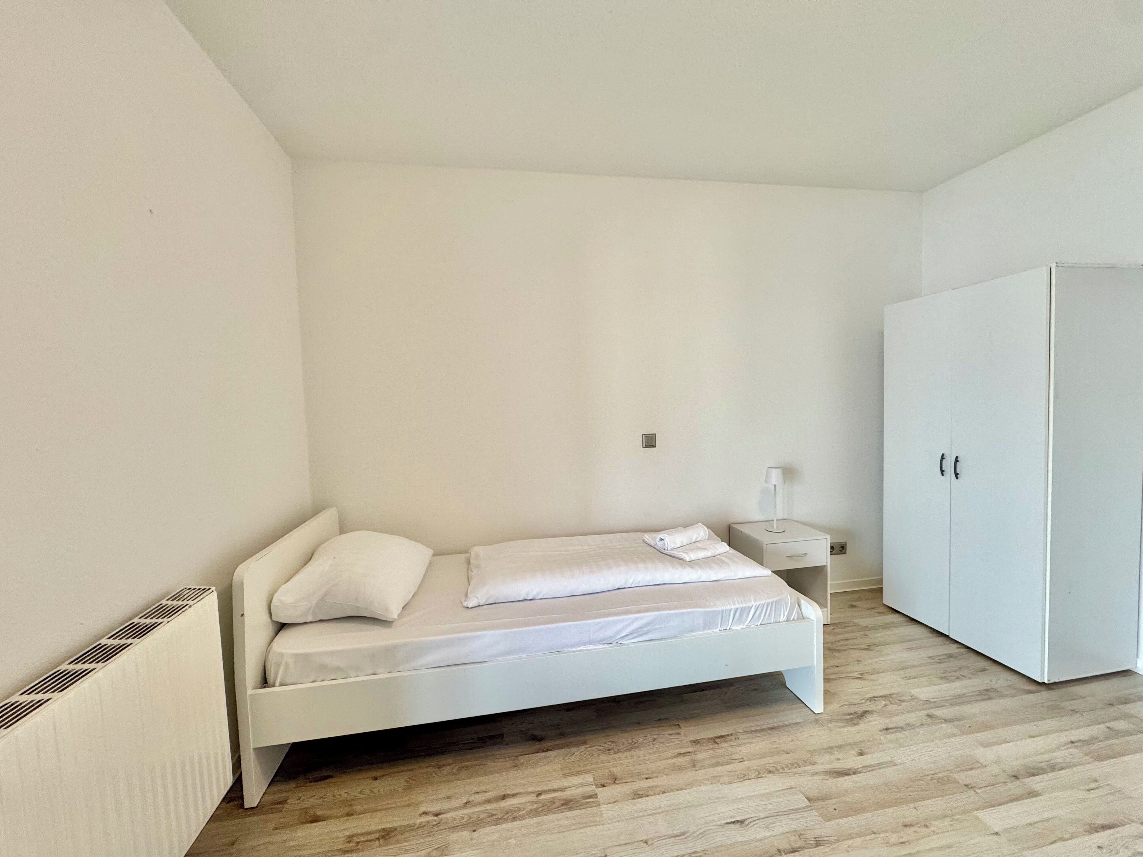 A small, white, single-bed room with a wooden floor, a white wardrobe, and a bedside table with a lamp.