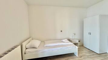 A small, white, single-bed room with a wooden floor, a white wardrobe, and a bedside table with a lamp.