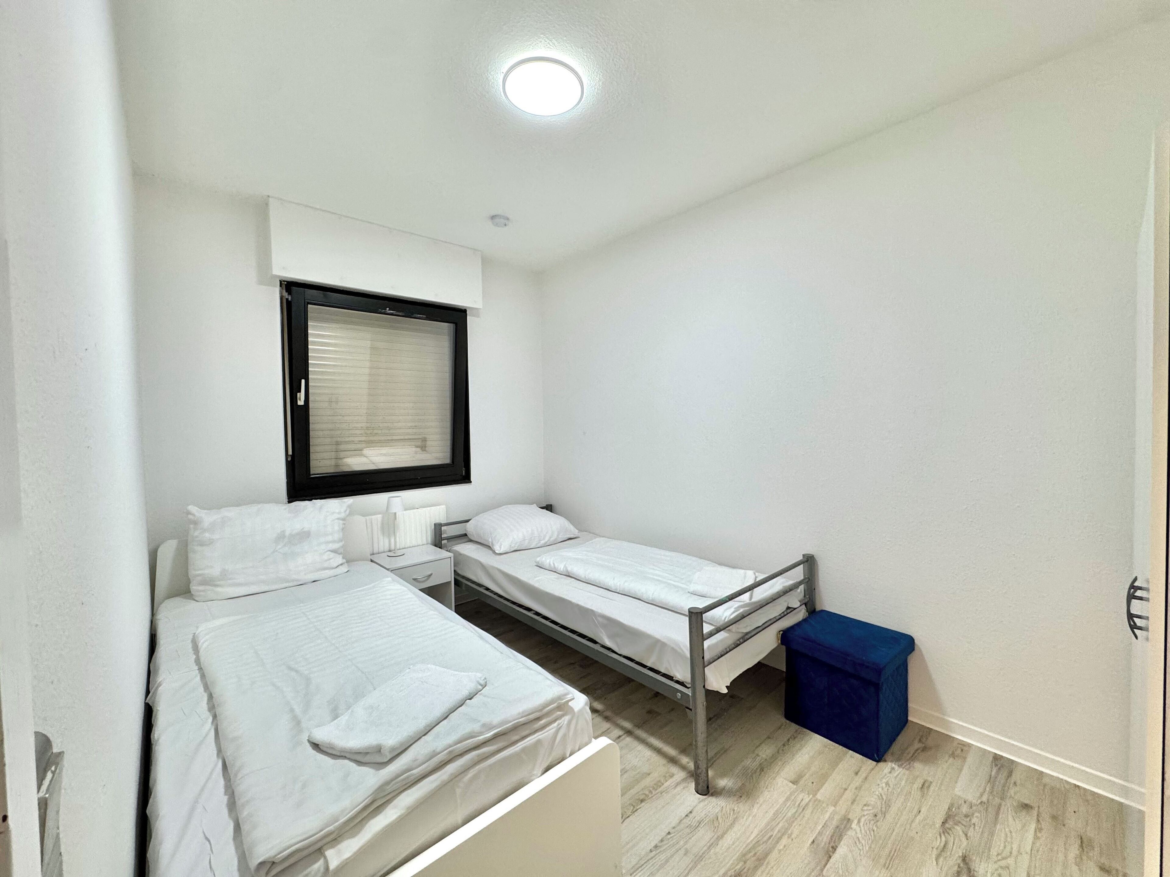 A small, clean room with two single beds, a window with blinds, a bedside table with a lamp, and a blue trash bin.
