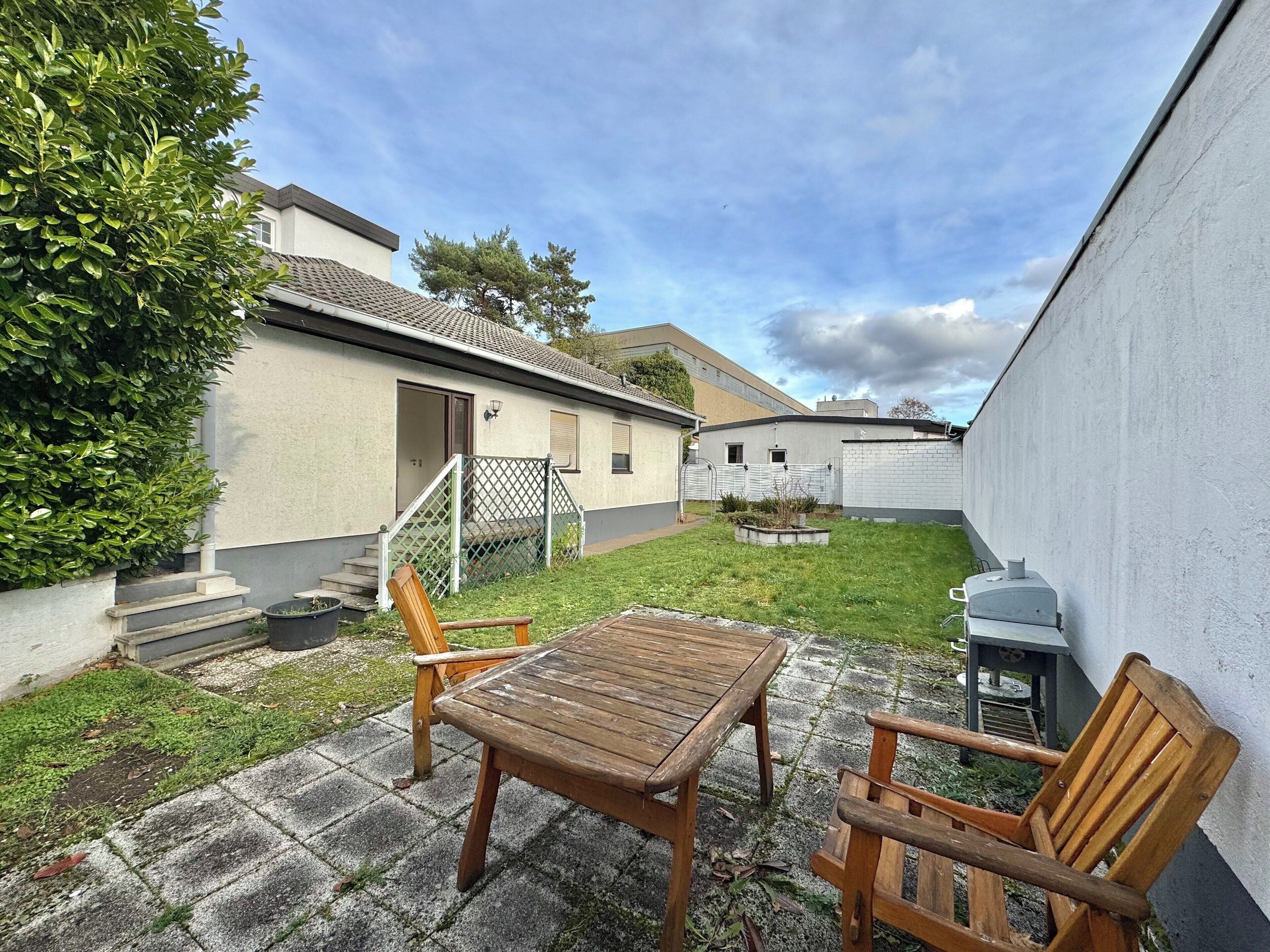 Un patio con mesa y sillas de madera, un pequeño jardín y un edificio con techo de tejas.