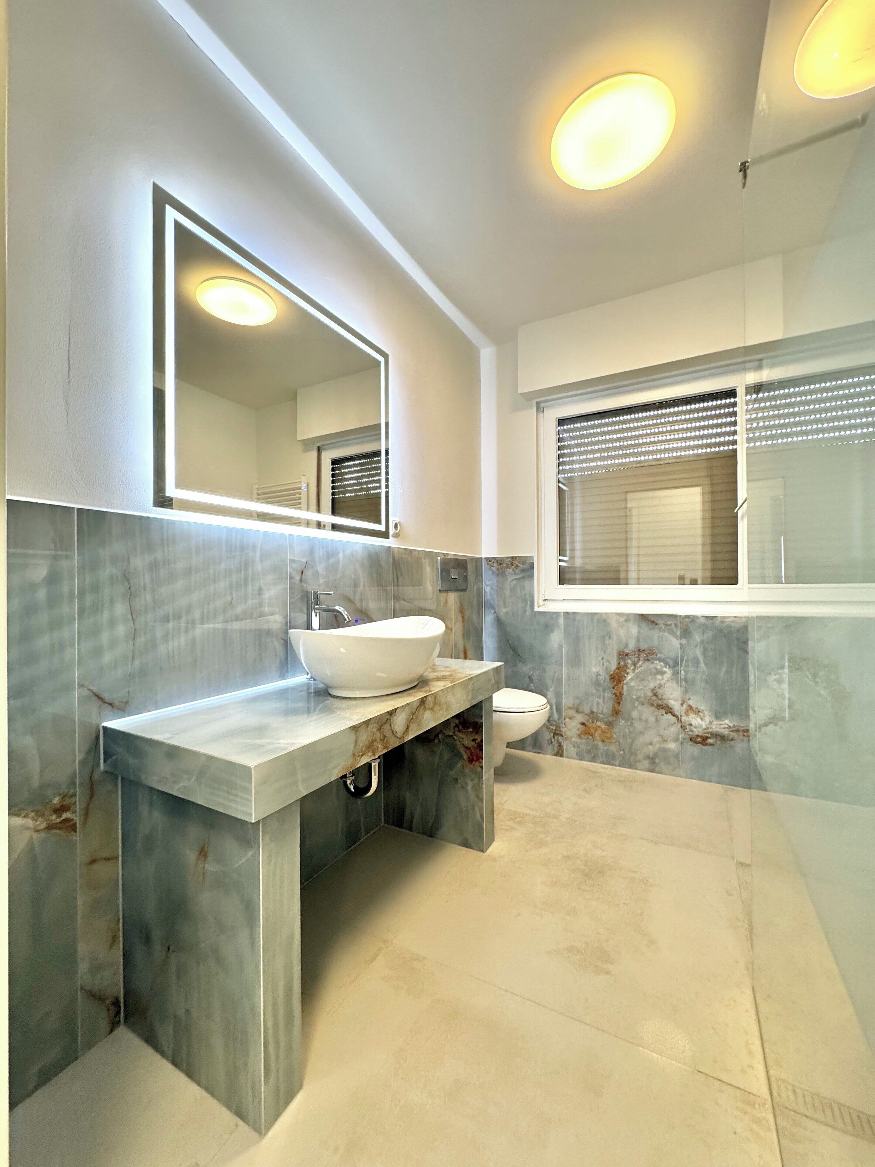 A modern bathroom with a marble vanity, a large mirror, a toilet, and a window with blinds.