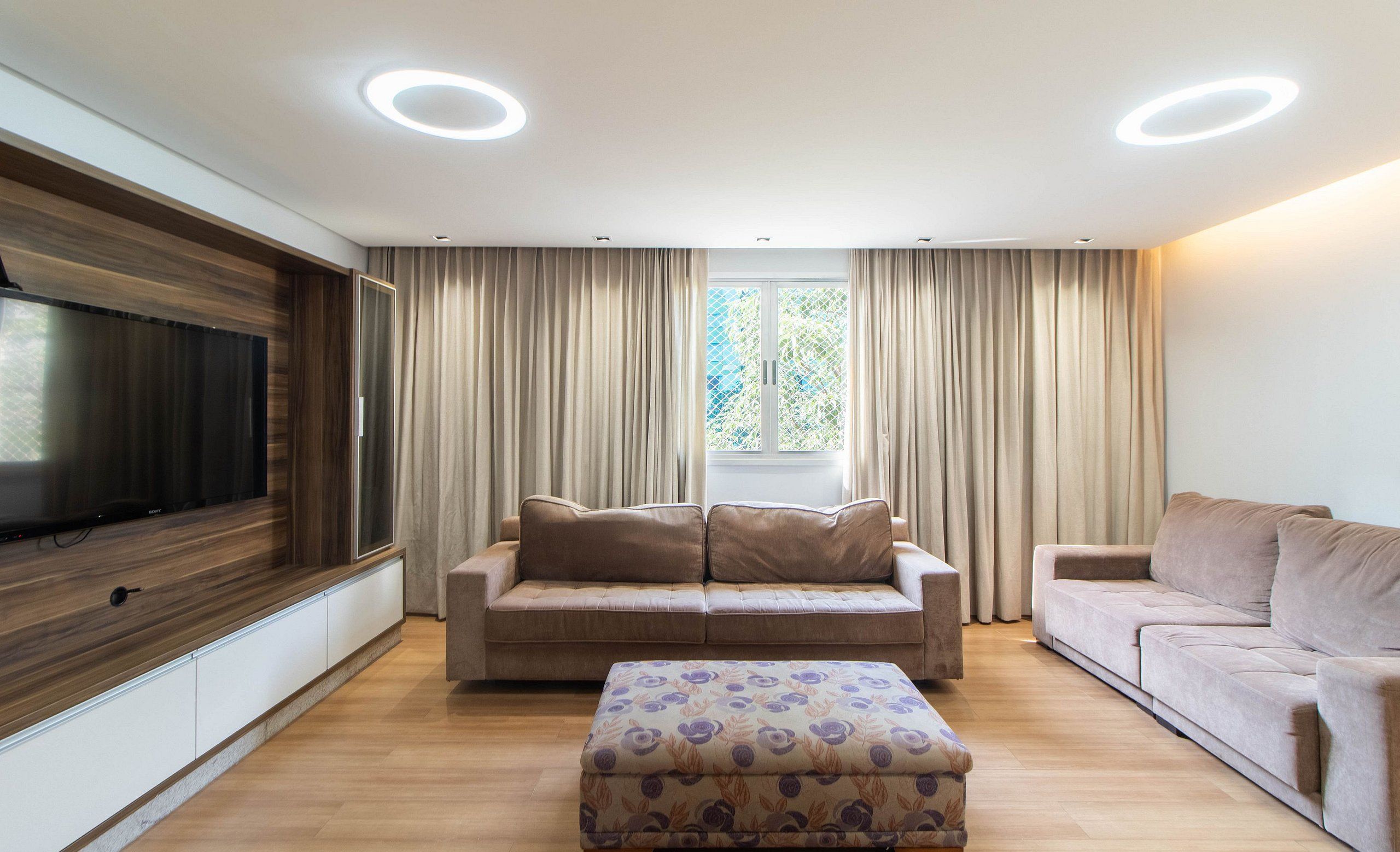 Una sala de estar moderna con un televisor de pantalla plana grande, dos sofás, un reposapiés estampado y un amplio ventanal con cortinas.
