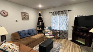 A living room with a brown sofa, a black ottoman, a bookshelf, a television, and a patterned curtain.