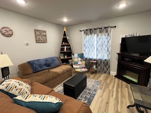 A living room with a brown sofa, a black ottoman, a bookshelf, a television, and a patterned curtain.