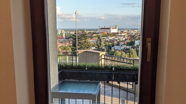 Ein Fenster mit Blick auf eine Stadtlandschaft, darunter ein schlossartiges Gebäude, ein Tisch und ein Geländer.