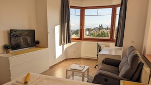 Ein modernes Hotelzimmer mit Bett, einer Couch, einem kleinen Tisch, einem Fernseher und einem Fenster mit Blick auf Gebäude und Bäume.