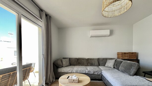 A modern living room with a grey sectional sofa, a round wooden coffee table, and a woven ceiling light fixture. - STAY Paradise Apartment (Protaras)