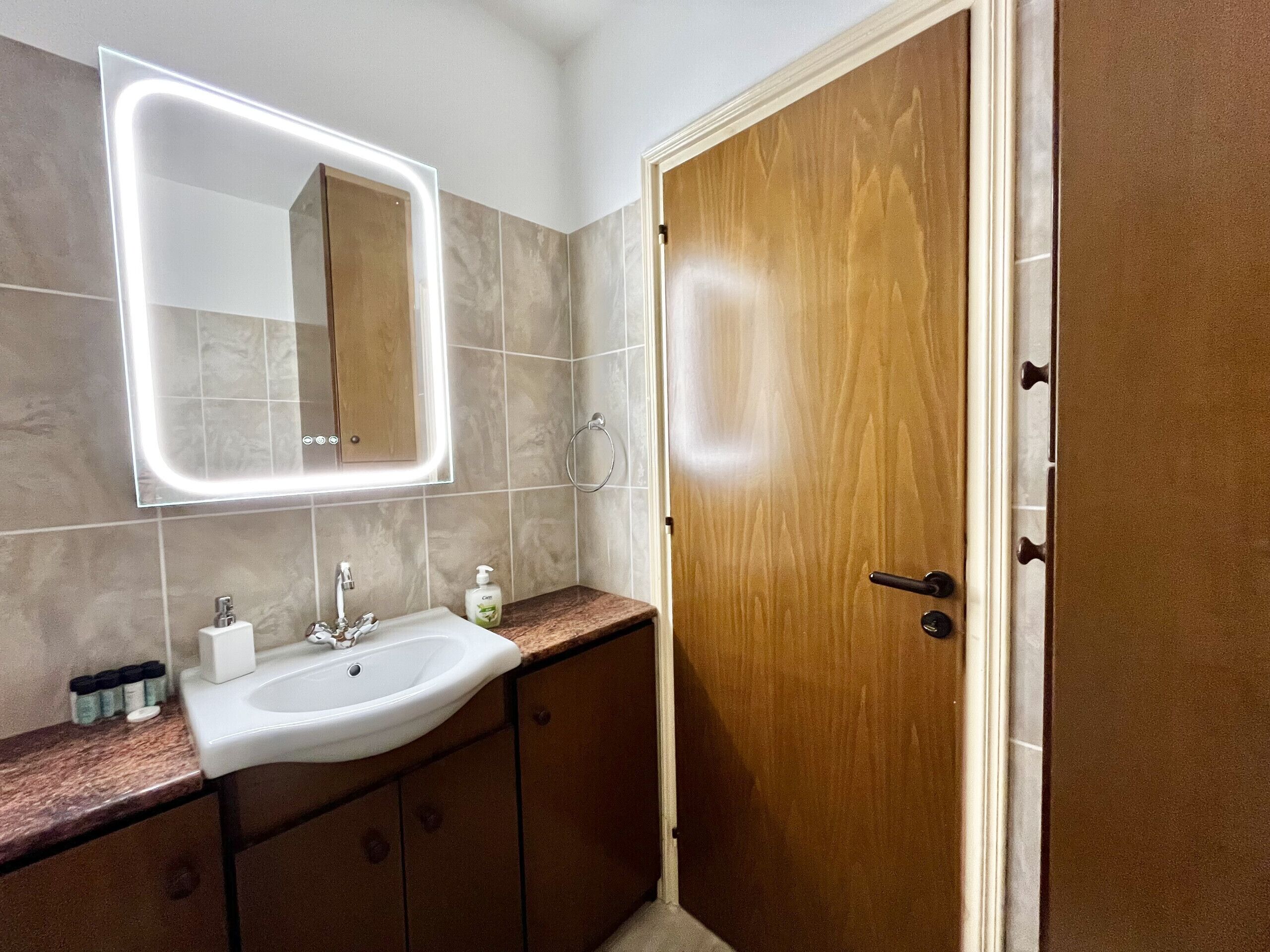 A bathroom with a white sink, a mirror with a light, and a wooden door.