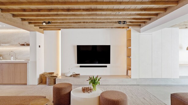 Una sala moderna con un televisor de pantalla plana montado en la pared, una mesa de centro redonda con objetos decorativos y otomanas marrones.