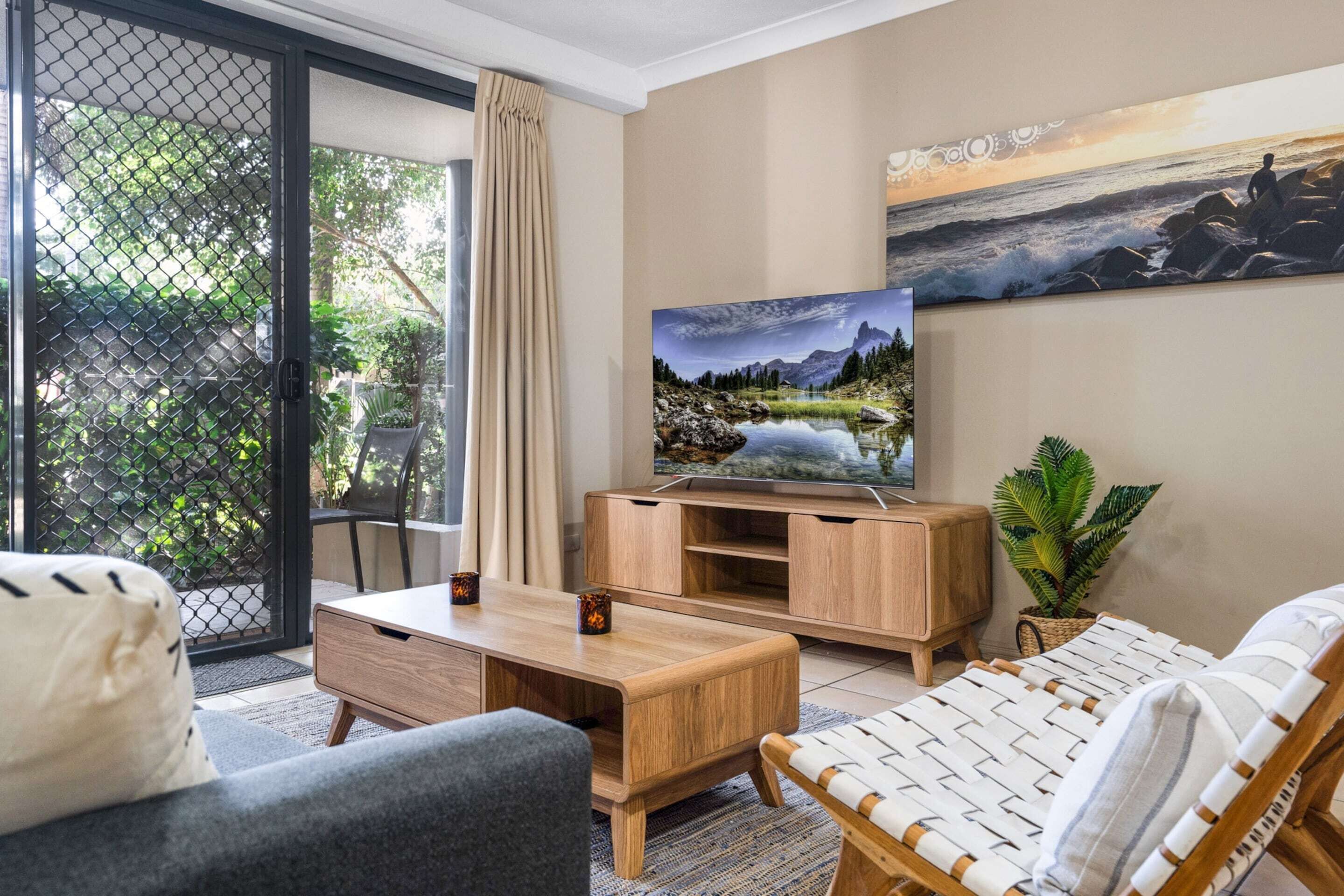 A modern living room with a wooden TV stand, a sofa, and a chair. There is a large window with a view of greenery outside.