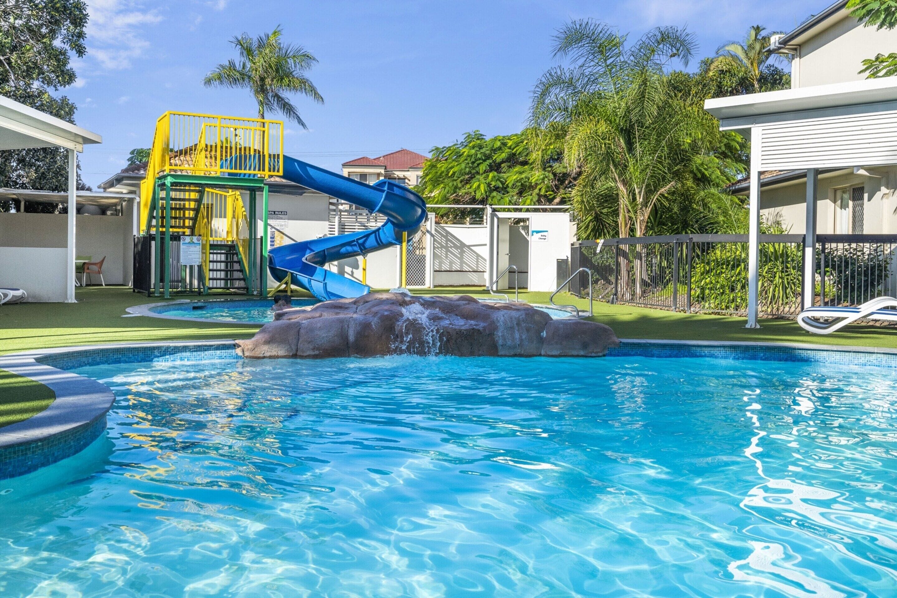Una piscina con un tobogán de agua azul, una formación rocosa y una zona cubierta con barandilla amarilla.