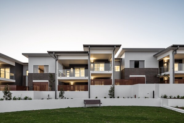 A modern multi-story residential building with balconies, a well-maintained lawn, and a bench in the foreground. - Pristine 3-bed Kiama Unit With Courtyard (Kiama)