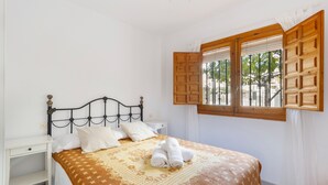Une chambre à coucher avec un lit, des tables de chevet blanches, une fenêtre avec des volets en bois et une vue sur la verdure à l’extérieur.