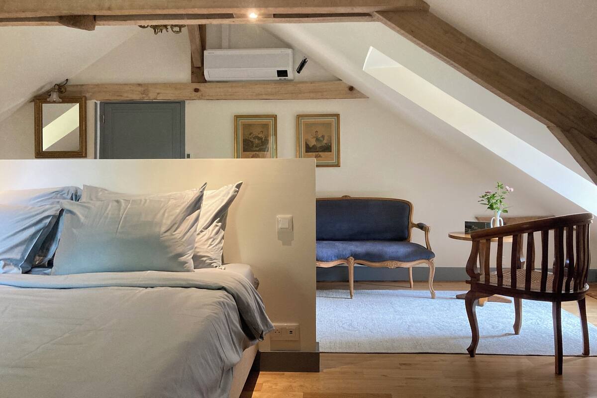An attic bedroom with a bed, a chair, and a table. There are framed pictures on the wall and a mirror above the bed.