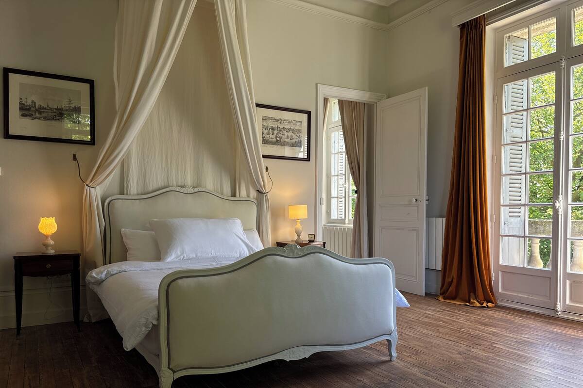 A bedroom with a four-poster bed, two bedside tables with lamps, a large window with curtains, and framed pictures on the wall.