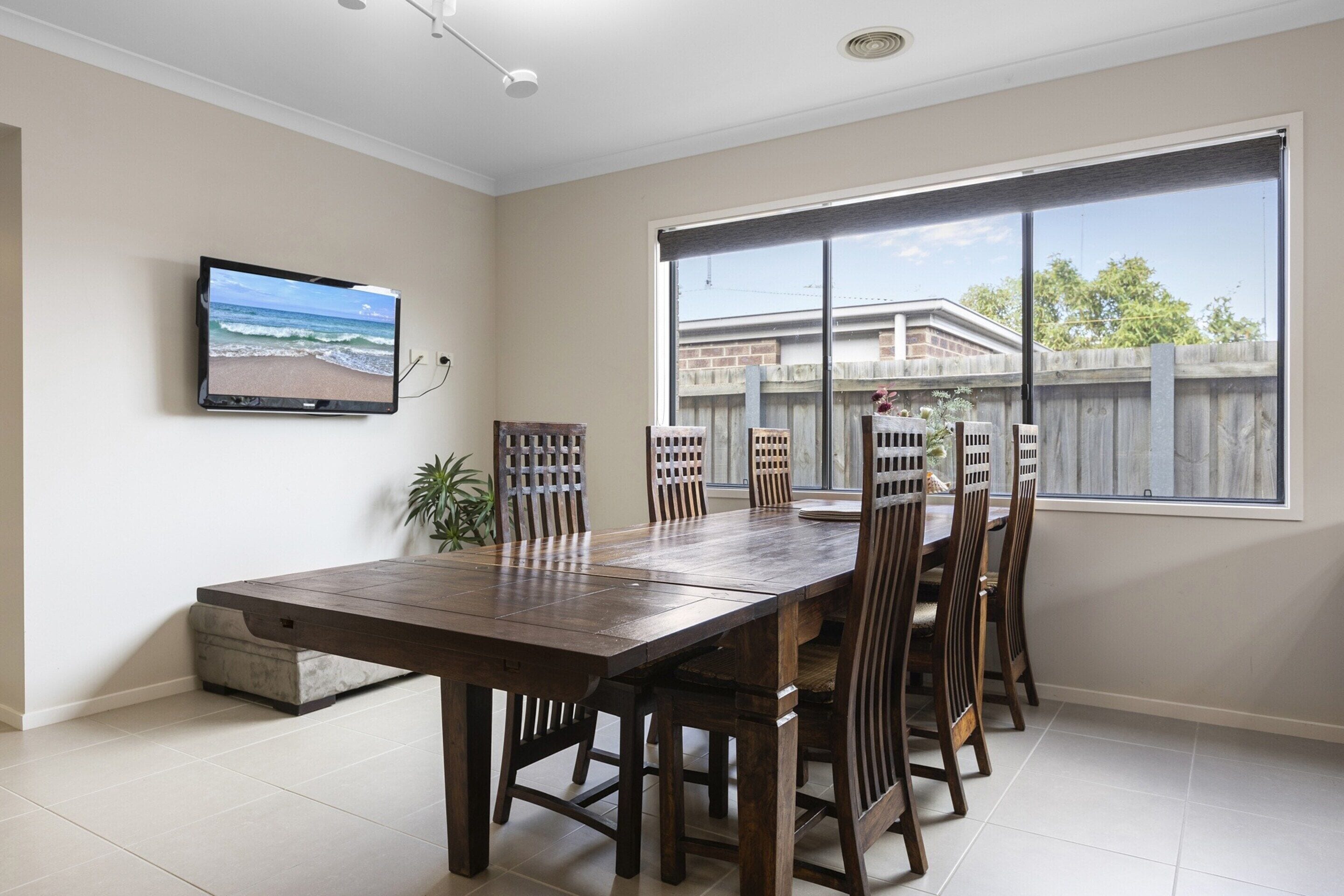 A dining area with a large table and chairs, a television on the wall, and a view of a backyard with a wooden fence.