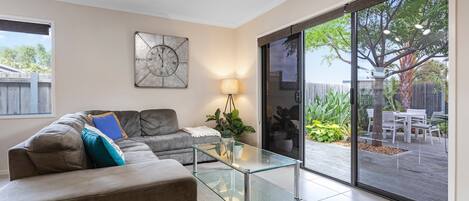 A modern living room with a glass coffee table, grey sofa, and a large window overlooking a garden.