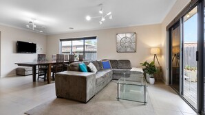 Ein modernes Wohnzimmer mit einer Eckcouch, einem Glastisch, einem Essbereich mit Tisch und Stühlen, einem an der Wand befestigten Flachbildfernseher und einer großen Schiebeglastür, die auf eine Außenpatio führt.