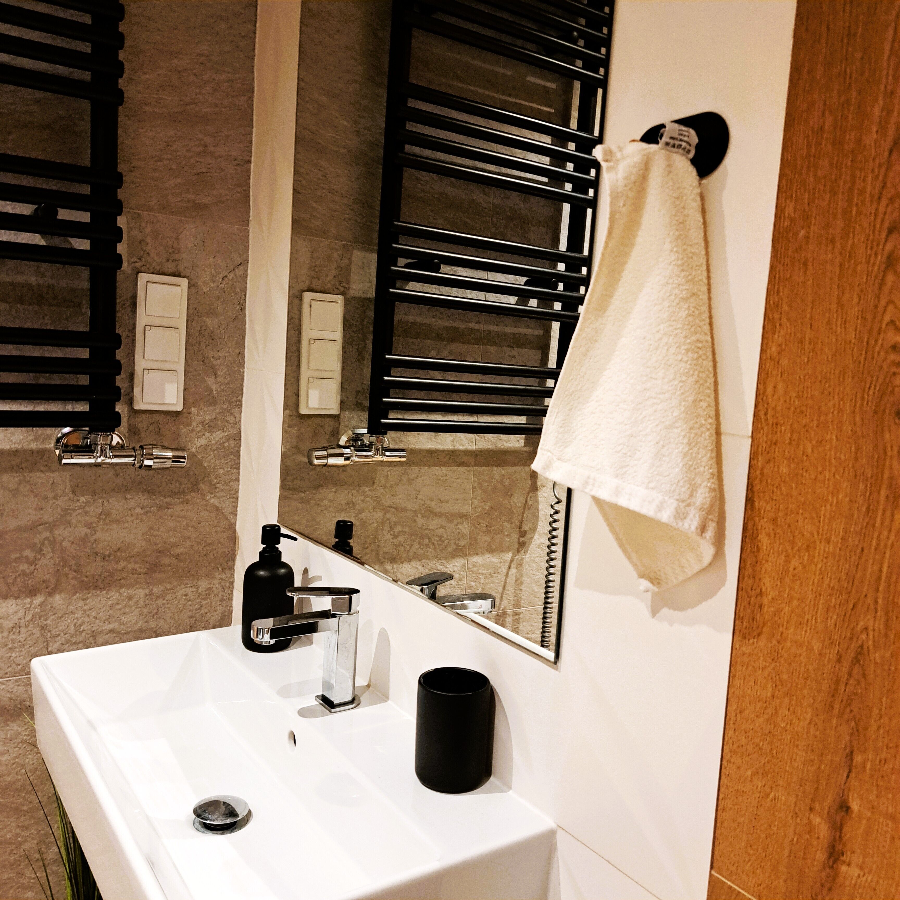 Ein Badezimmer mit einem weißen Waschbecken, einem schwarzen Seifenspender und einem Spiegel, der einen Handtuchhalter reflektiert.