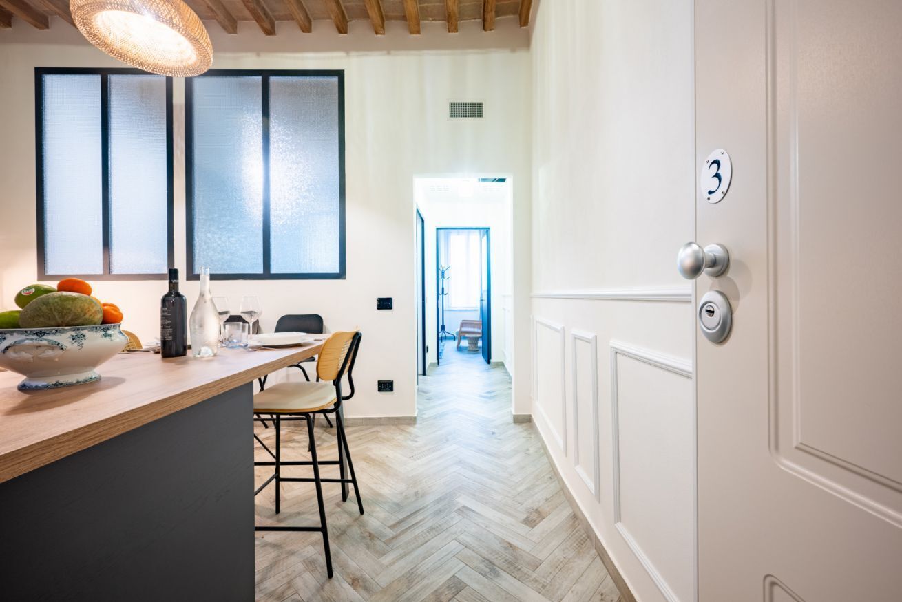 Une cuisine avec un plan de travail en bois, un saladier rempli de fruits, une lampe suspendue et une porte portant un numéro.