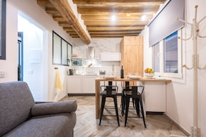 A compact kitchen with a wooden ceiling, a dining area with two black chairs, and a grey sofa.